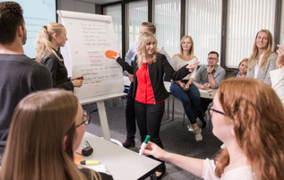 Eine Dozentin steht mit jungen Menschen, die an einem Kurs teilnehmen, vor einem Flipchart und klebt einen Zettel mit Inhalten auf.